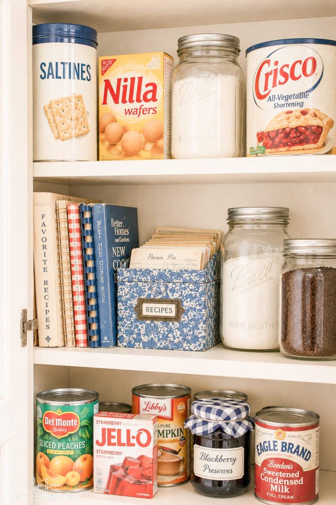 Vintage kitchen cabinet stocked with Saltines, vanilla wafers, Crisco, canned peaches, Jell-O, preserves, and old cookbooks.