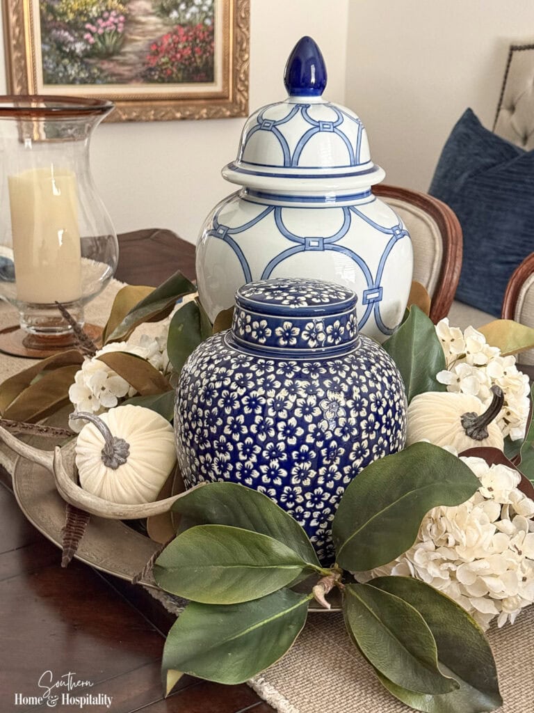 Fall centerpiece with magnolia leaves, blue and white pottery, dried hydrangeas, white pumpkin, antler, and pheasant feather