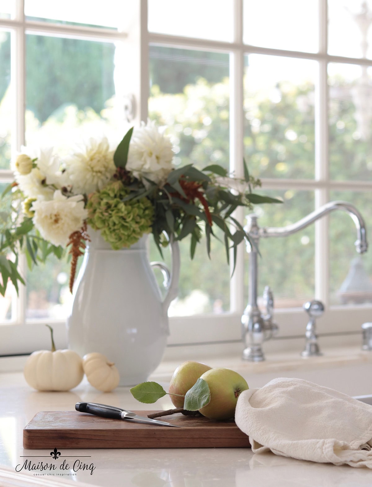 Green and white flowers in a white pitcher with pumpkins next to a kitchen window