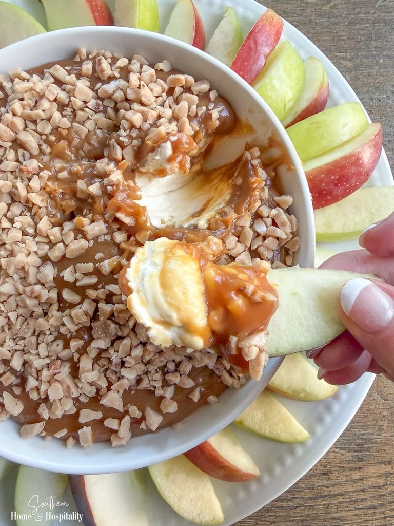 Hand holding an apple slice covered with cream cheese, caramel, and toffee bits above a bowl of caramel apple dip.