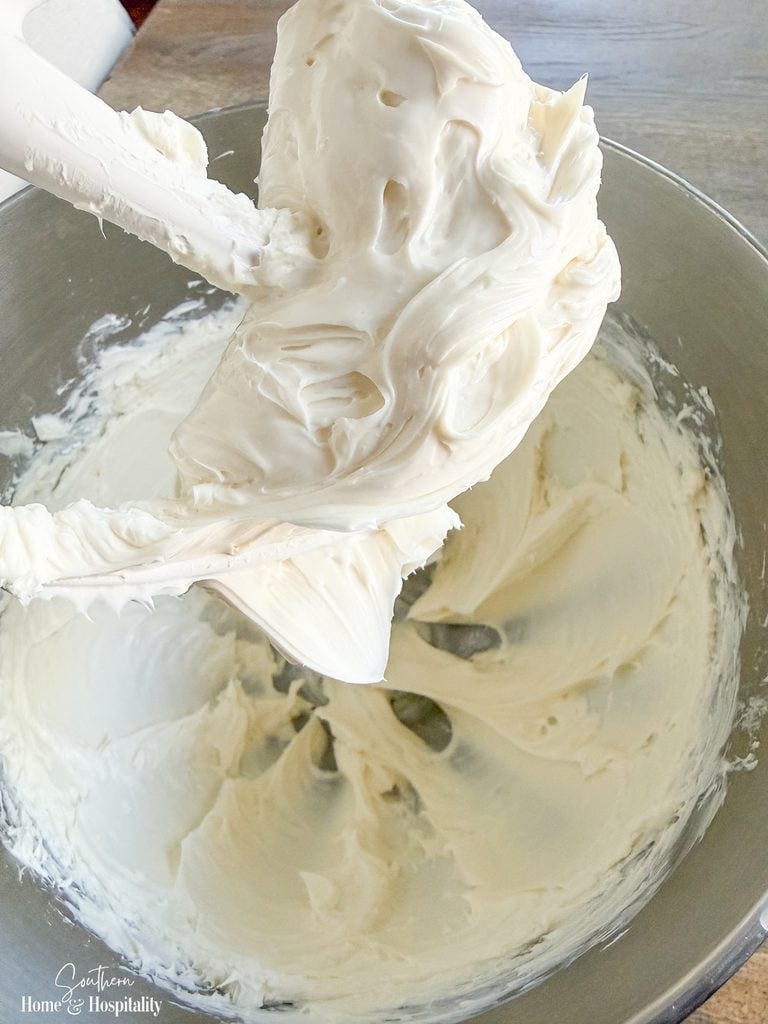 Electric mixer beaters lifting smooth, fluffy cream cheese mixture from a glass mixing bowl.