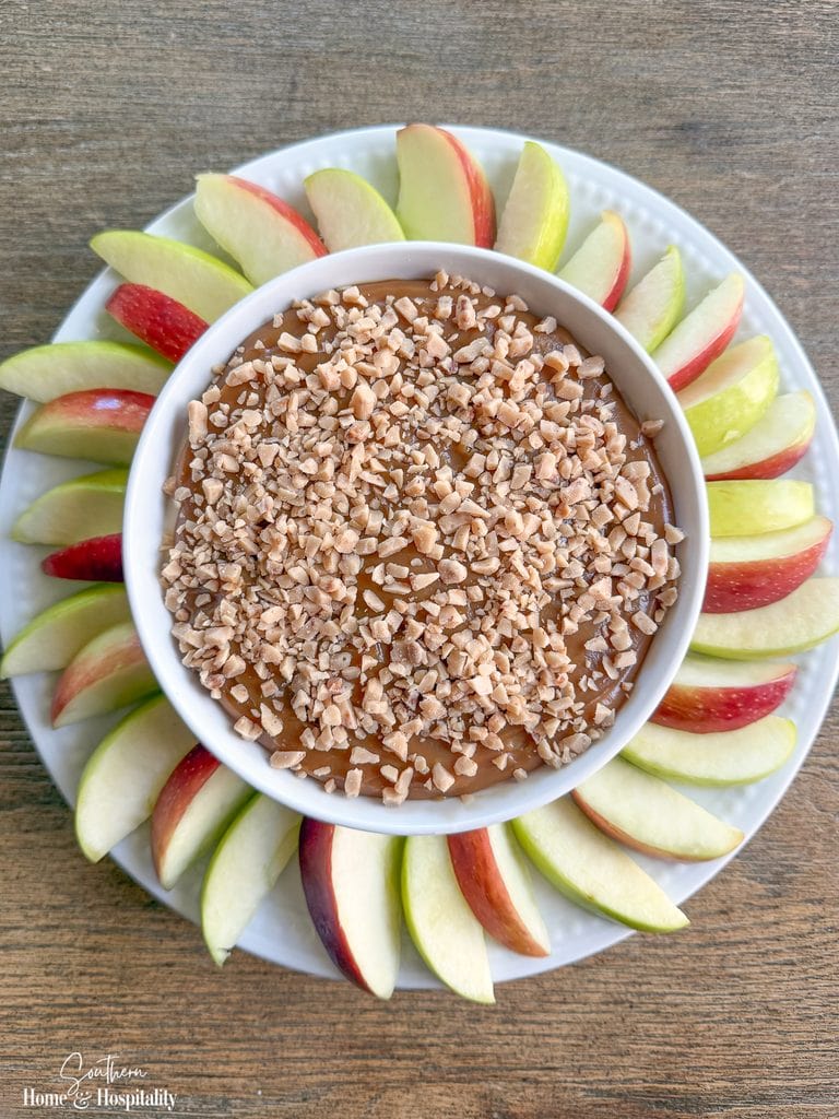 Finished caramel apple dip in a white bowl surrounded by sliced red and green apples on a serving platter.