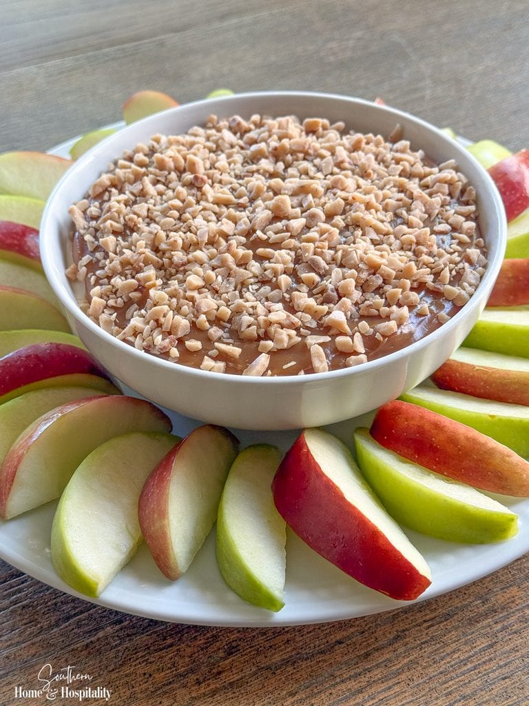 Angled view of a bowl of caramel apple dip topped with toffee bits and served with red and green apple slices.