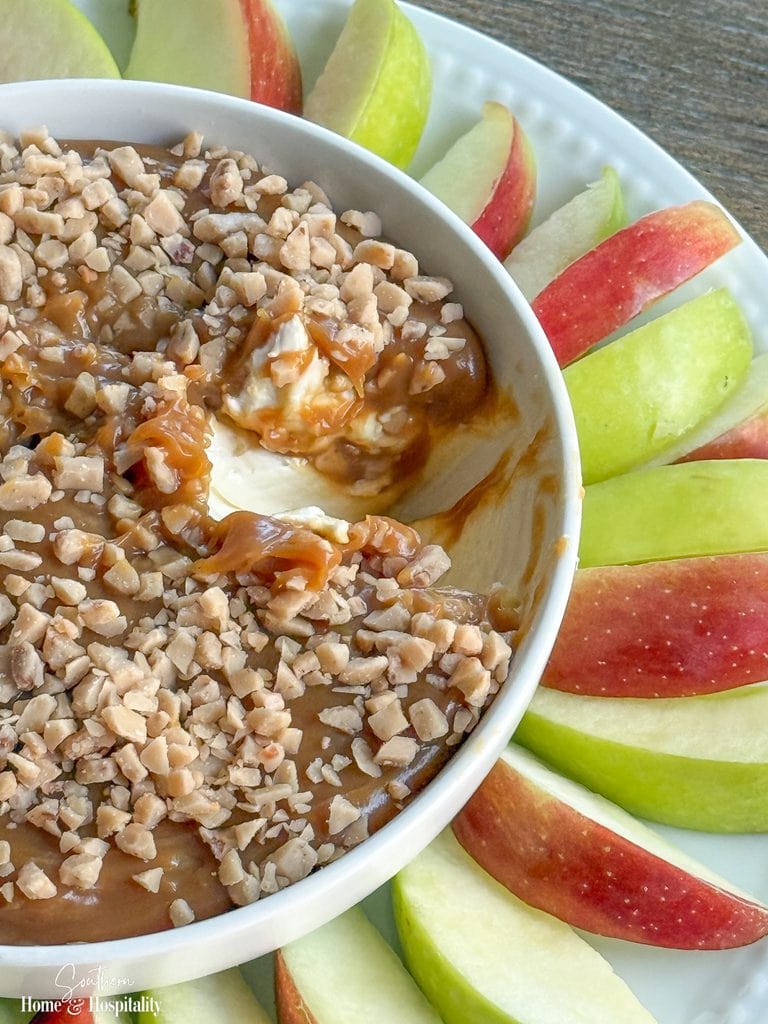 Close-up of caramel apple dip with a scoop removed, showing the cream cheese layer beneath the caramel and toffee topping.