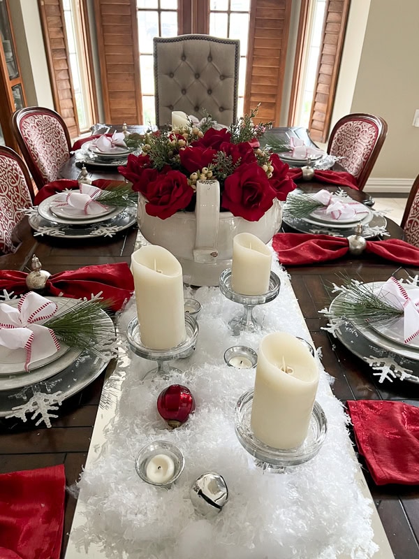 Long Christmas dining table set with red and white decorations, a snowy greenery centerpiece, candles, and festive place settings